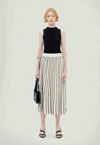 Top sans manches noir à côtes associé à une jupe blanche mi-longue ornée de rayures verticales noires. La mannequin tient un petit sac à main noir.