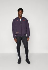 Herrbomberjacka i mörklila nylon med dragkedja framtill, ribbade muddar och broderade detaljer. Matchas med svarta byxor med vita ränder.