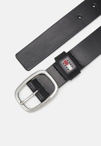 La ceinture en cuir noir est dotée d'une boucle en métal argenté arrondie et d'un patch de logo de marque en rouge, blanc et bleu sur la lanière de la ceinture.