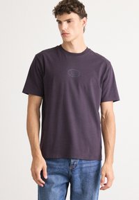 Lila pamut póló rövid ujjakkal, kerek nyakkivágással és domború logóval a mellkasán. Kék farmernadrággal párosítva.