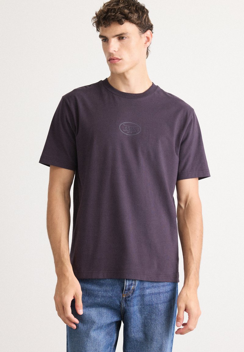 Lila pamut póló rövid ujjakkal, kerek nyakkivágással és domború logóval a mellkasán. Kék farmernadrággal párosítva.