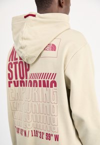 Sudadera beige con un gran diseño gráfico en texto burdeos que dice "NUNCA DEJES DE EXPLORAR" con coordenadas en la sección inferior.
