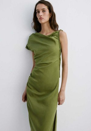 Mujer con cabello largo y oscuro, vestida con un vestido sin mangas de color verde oliva con tela drapeada, de pie frente a una pared blanca.