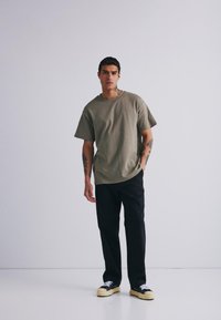 Tricou din bumbac de culoare verde măsliniu, cu mâneci scurte, decolteu rotund și croială lejeră, asortat cu pantaloni negri și adidași negri cu talpă albă.