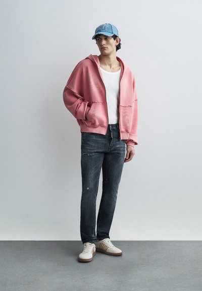 Ροζ φαρδιά φούτερ με μπροστινές τσέπες, φορεμένο πάνω από λευκή αμάνικη μπλούζα· συνδυασμένο με σκούρο τζιν και ανοιχτόχρωμα αθλητικά παπούτσια με μπεζ λεπτομέρειες.