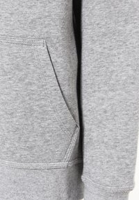 Grijze sweatstof met een getextureerd, zachte uitstraling. Voorzien van een kangoeroezak en geribbelde manchetten, wat een ontspannen ontwerp benadrukt.