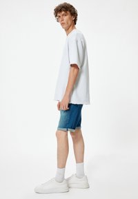 Camiseta de algodón gris de corte oversized con mangas deshilachadas, combinada con unos pantalones cortos de mezclilla que presentan un degradado de oscuro a claro. Zapatillas blancas y calcetines.