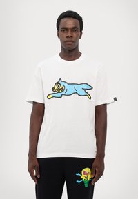 Hvid bomulds T-shirt med en blå, tegneserieagtig kat med et gult ansigt og skarpe tænder. Korte ærmer, rund halsudskæring, afslappet pasform.