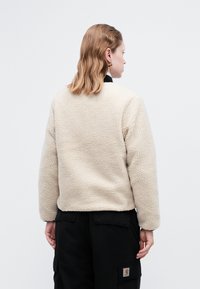 Vaaleanbeige fleece-takki, jossa on teksturoitu kangas, pyöreät olkapäät, pitkät hihat ja mustat yksityiskohdat kauluksessa, yhdistettynä tummiin cargo-housuihin.
