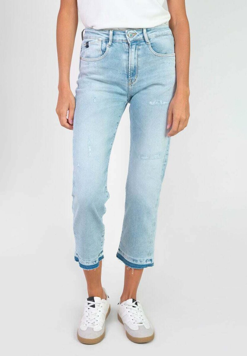 Jean cropped en denim bleu clair avec un ourlet effiloché, présentant des détails déchirés et un design taille haute, associés à des baskets blanches.