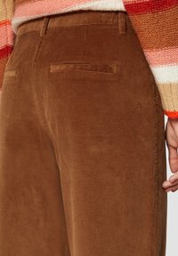 Bruine corduroy broek met verticale ribbels, voorzien van twee achterzakken en een lichte structuur, gecombineerd met een multicolor gestreept sweater.