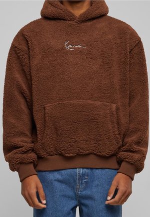 Person trägt einen braunen Fleece-Hoodie mit Fronttasche und Logo, kombiniert mit blauen Jeans vor einfachem Hintergrund.