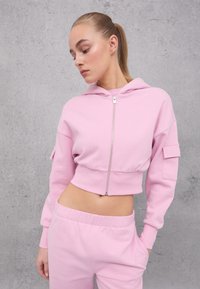 Felpa corta color rosa chiaro, realizzata in materiale morbido, presenta una zip frontale, cappuccio con cordino e tasche laterali. Abbinata a pantaloni coordinati.