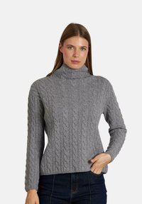 Maglione grigio a maglia intrecciata con un alto collo, caratterizzato da motivi testurizzati e maniche lunghe, abbinato a jeans in denim scuri.