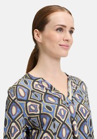 Langärmlige Bluse in Blau mit einem geometrischen Muster aus Rauten und Ovalen in Rot, Creme und Schwarz. Mit angeschnittenem Kragen und weicher Textur.