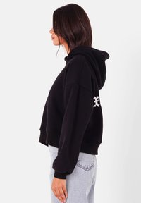 Svart croppad hoodie med huva, med en vit logotyp på vänster ärm. Tillverkad av mjukt material med en avslappnad passform och ribbade muddar.