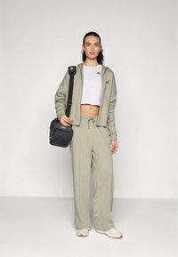 Ljusgrå sportoverall med jacka med dragkedja och byxor med dragsko. Kompletteras med en svart messenger-väska och vita sneakers.