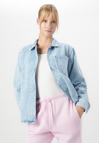 Jeune femme aux cheveux blonds portant une veste en denim bleu clair, un haut à côtes blanc et un pantalon à cordon rose clair, posant sur fond blanc.