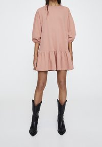 Rosa Sweatshirt-Kleid mit Puffärmeln, einem Rüschensaum und einem runden Halsausschnitt, kombiniert mit schwarzen Cowboystiefeln. Glatte Stofftextur.