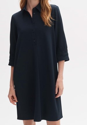 Woman wearing a loose-fitting, knee-length black dress with three-quarter sleeves and a buttoned collar.