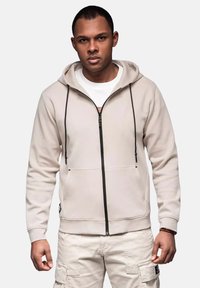 Beige zip-hoodie med sort lynlås og snøreb�ånd, med en frontlomme og ribbede manchetter. Båret over en hvid skjorte og lyse bukser.