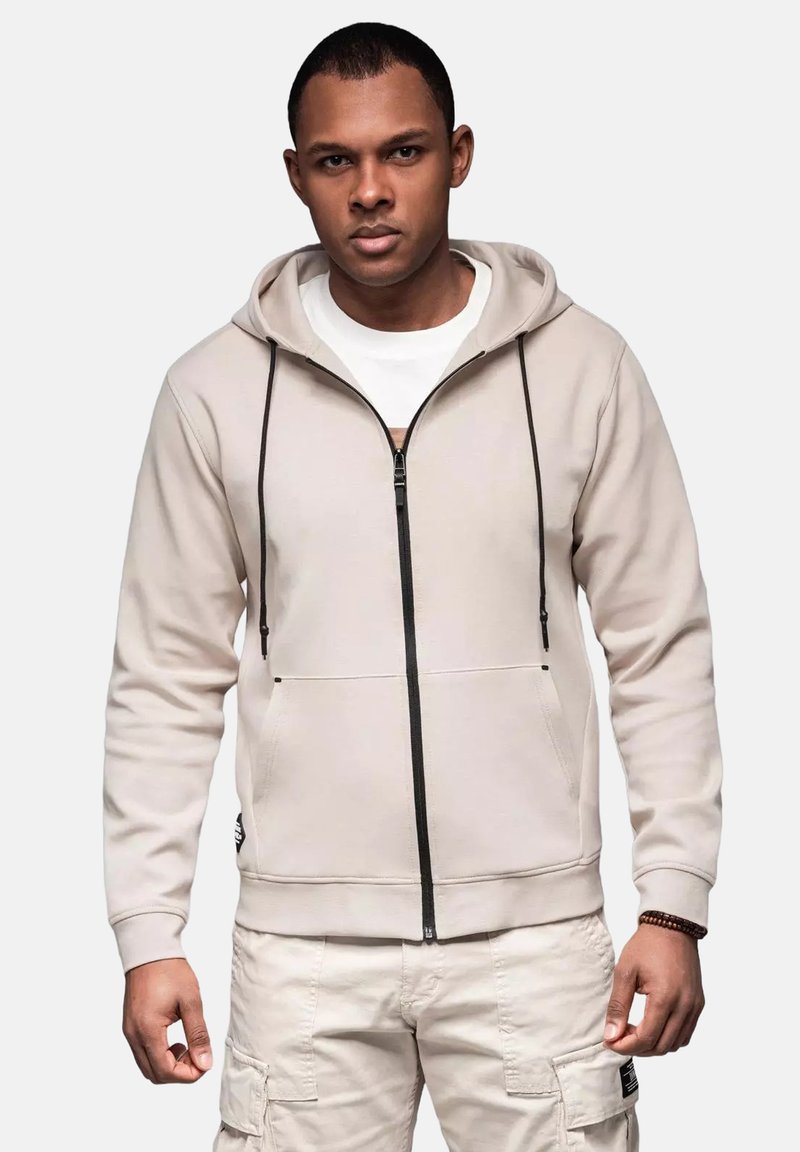 Beige zip-hoodie med sort lynlås og snørebånd, med en frontlomme og ribbede manchetter. Båret over en hvid skjorte og lyse bukser.