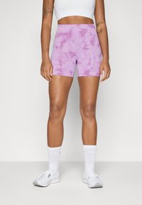 Shorts de sport à motifs tie-dye violets avec une taille haute, fabriqués en tissu extensible, associés à des chaussures blanches et des chaussettes jusqu'aux chevilles.