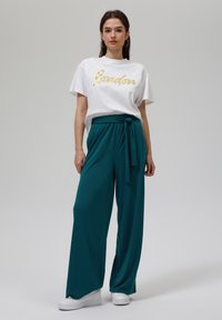 T-shirt blanc avec "Londres" en écriture dorée, associé à un pantalon ample teal avec une ceinture à nouer à la taille. Des baskets blanches complètent la tenue.