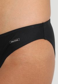 Črne bikini hlačke z gladko teksturo, z majhno srebrno oznako na strani. Preprost dizajn, oblikovane za minimalno pokritost.