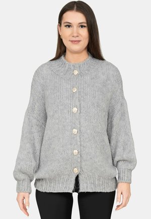 Vrouw die een lichtgrijze grofgebreide cardigan draagt met grote crèmekleurige knopen, staand met ontspannen armen tegen een effen achtergrond.