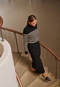 Un pull rayé noir et beige avec un col haut, associé à une jupe noire ajustée, descendant un escalier circulaire avec une rampe en cuivre.