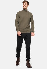 Zeleni pletivi pulover s turtleneck izrezom i cik-cak uzorkom, kombiniran s crnim uskim trapericama i smeškim čizmama sa vezicama. Glatka tekstura, rebrasti manžeti.