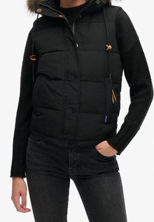 Persona que lleva un chaleco negro acolchado con capucha con borde de piel sobre un suéter de punto negro, con una mano en el bolsillo de unos vaqueros oscuros.