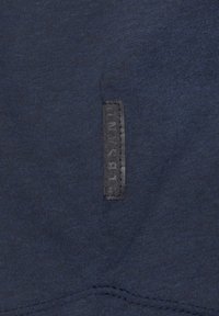 Dunkelblauer Sweatshirtstoff mit einer strukturierten Oberfläche und einem rechteckigen, schwarzen Label, das an der Seite aufgenäht ist und den Markennamen zeigt.
