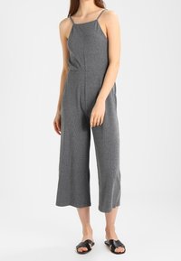 Mono gris con tirantes finos, un corpiño ajustado y piernas amplias cortas; hecho de un tejido suave y texturizado; combinado con sandalias negras.