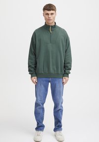 Sudadera verde con cremallera y cuello alto, puños y dobladillo acanalados, combinada con unos jeans azul claros y zapatillas blancas. Tejido suave y texturizado.