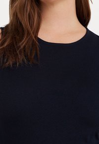 Námořnická modrá bezrukávová blůza s kulatým výstřihem, vyrobená z hladkého materiálu. Viditelné ploché švy kolem výstřihu a oblasti ramen.