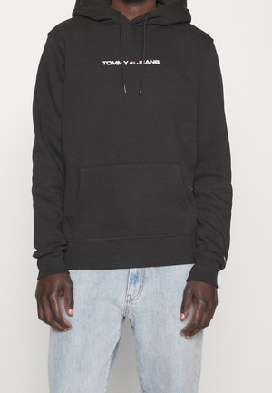 Mann trägt schwarzen Tommy Jeans Hoodie mit Fronttasche und hellblauen Jeans vor schlichten Hintergrund.