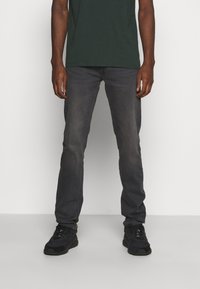 Jeans estrechos de color gris oscuro con desvanecimiento visible, hechos de tela de mezclilla. Combinados con zapatillas negras y una camiseta verde. Diseño simple, sin patrones.