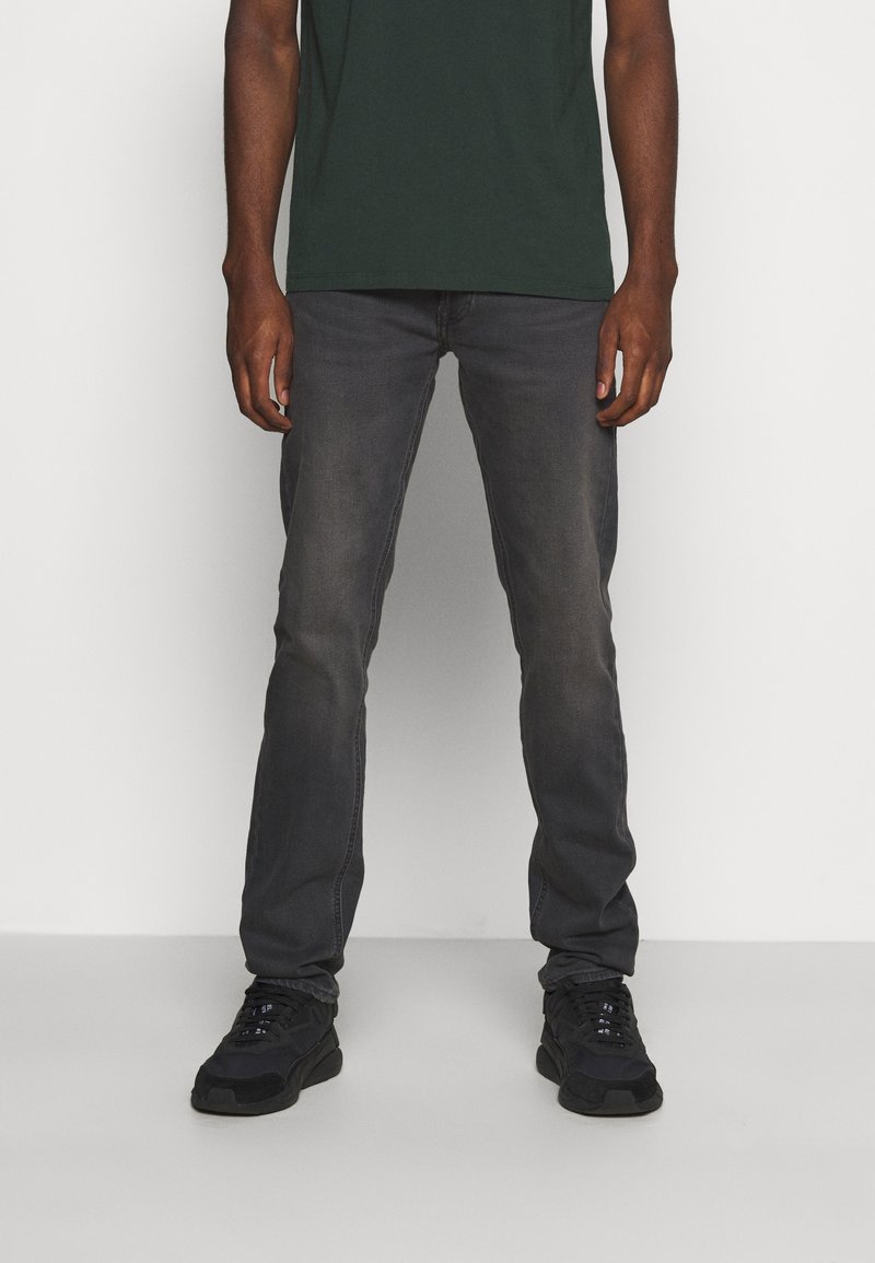 Jeans estrechos de color gris oscuro con desvanecimiento visible, hechos de tela de mezclilla. Combinados con zapatillas negras y una camiseta verde. Diseño simple, sin patrones.