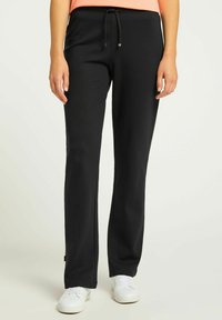 Zwarte sweatpants met een relaxte pasvorm, elastische tailleband en trekkoord. De stof lijkt glad en zacht. Gecombineerd met witte sneakers.