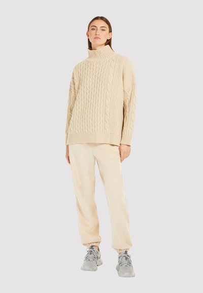 Suéter de punto beige con cuello alto y patrones de trenzado, combinado con joggers beige claros y zapatillas grises. Textura suave en toda la prenda.