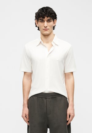 Jeune homme aux cheveux courts, bouclés et foncés, portant une chemise blanche à manches courtes avec boutons et un pantalon gris foncé, faisant face à l'avant sur un fond uni.