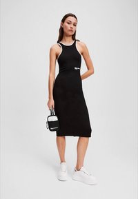 Černé šaty bez rukávů s přiléhavým střihem, bílé akcenty na výstřihu, doplněné bílými platformovými teniskami a malou černou kabelkou.