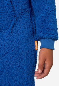 Otroška roka ob modrem puhastem flisu s pomarančno oznako z imenom "OPPOSUITS" na beli podlagi.