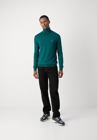 Pull à col roulé vert, coupe ajustée, ourlet et poignets côtelés, associé à un jean noir et des chaussures de sport multicolores.