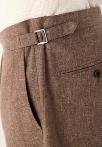 Pantalones marrones a medida hechos de una tela texturizada, con una hebilla plateada en la cintura, pliegues y un bolsillo trasero con botón.