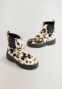 Botine cu imprimeu de vacă, dotate cu o pană elastică neagră, detalii cu cataramă și o talpă groasă neagră. Prezintă o suprafață texturată și accente contrastante.