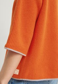 Jersey de punto naranja con mangas cortas y un patrón texturizado, con un ribete blanco en el dobladillo inferior y en los puños, y una pequeña etiqueta de la marca.