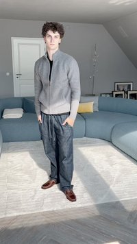 Pilkos užtrauktuko megztinis, juoda marškiniai, laisvi mėlyni džinsai ir rudi batai. Atsipalaidavęs stilius su šoninėmis kišenėmis, stovintis ant šviesaus kilimo.